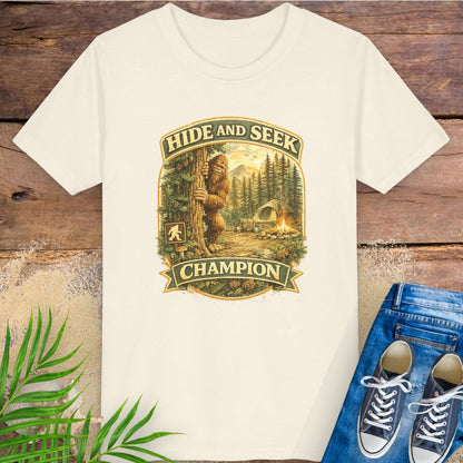 Light beige t-shirt with 'Hide and Seek Champion' graphic with Bigfoot peeking around a tree in a campsite.