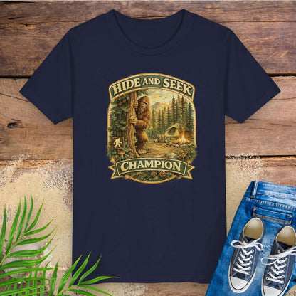 Navy blue t-shirt with 'Hide and Seek Champion' graphic with Bigfoot peeking around a tree in a campsite.