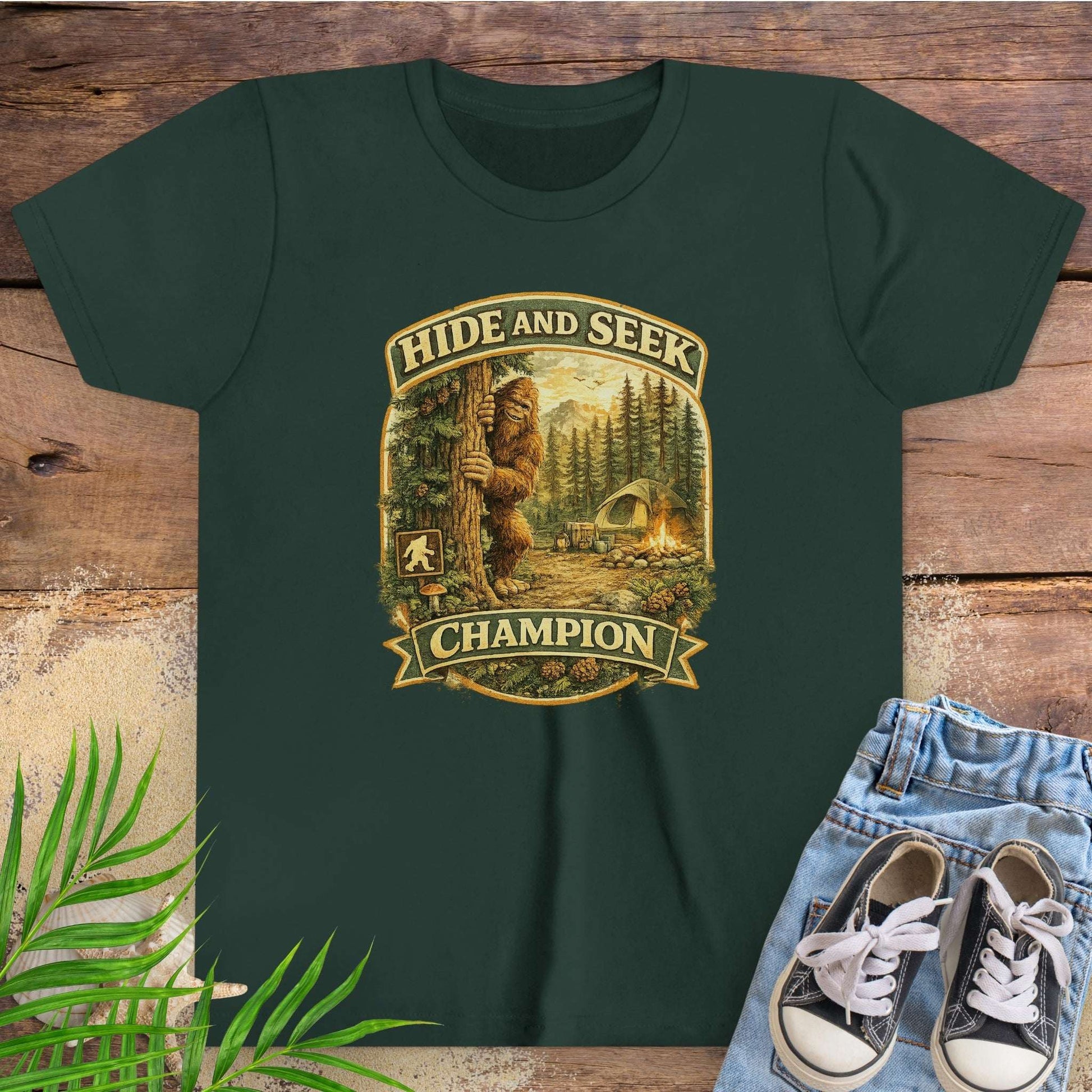 Green t-shirt with 'Hide and Seek Champion' graphic with Bigfoot peeking around a tree in a campsite.