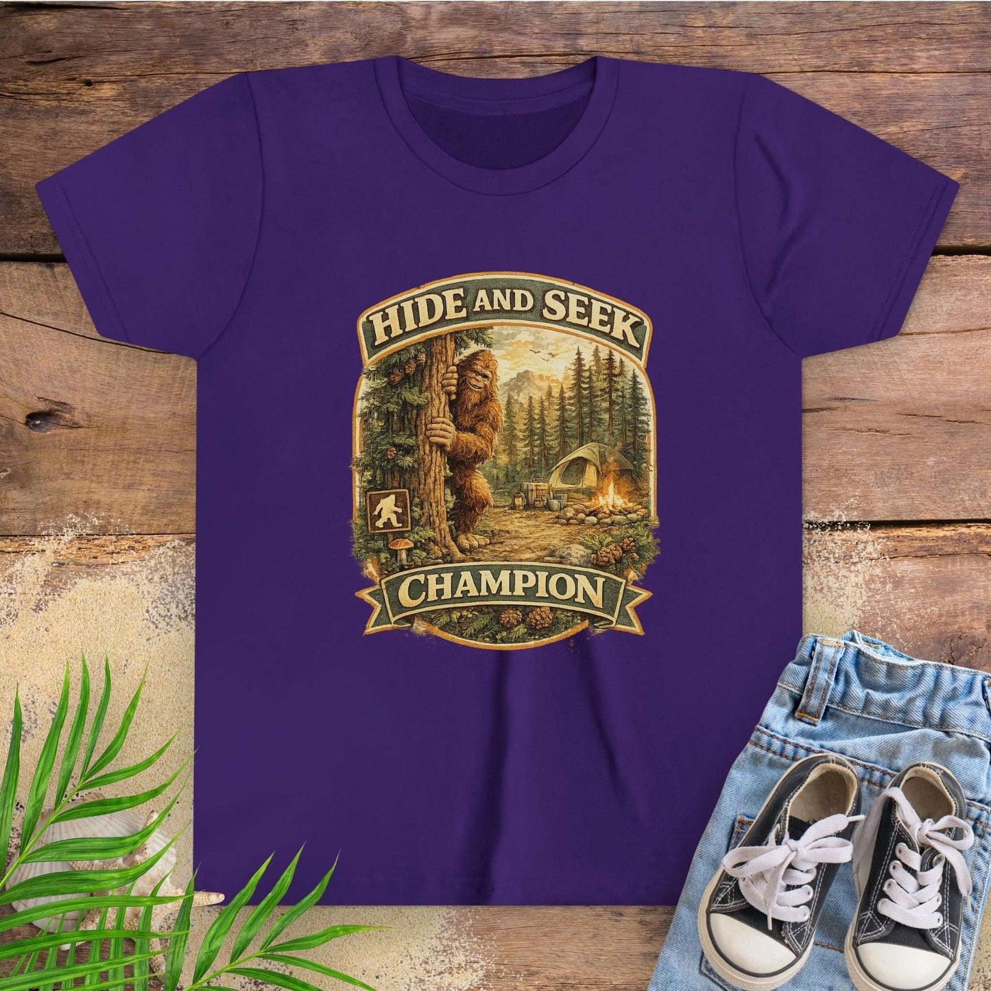 Purple t-shirt with 'Hide and Seek Champion' graphic with Bigfoot peeking around a tree in a campsite.