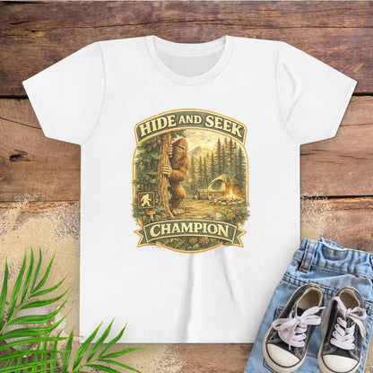 White t-shirt with 'Hide and Seek Champion' graphic on a wooden surface with sneakers and leaves.