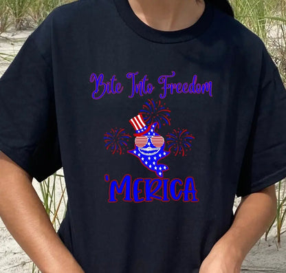 A Woman wearing a black t-shirt with a patriotic shark with red and white striped glasses, a patriotic top hat with fireworks around that says Bite Into Freedom "merica.