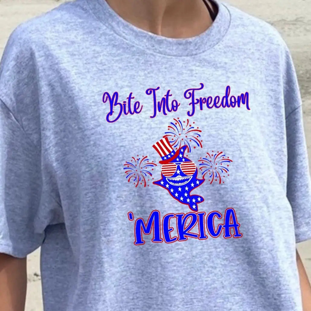 A Woman wearing a gray t-shirt with a patriotic shark with red and white striped glasses, a patriotic top hat with fireworks around that says Bite Into Freedom "merica.