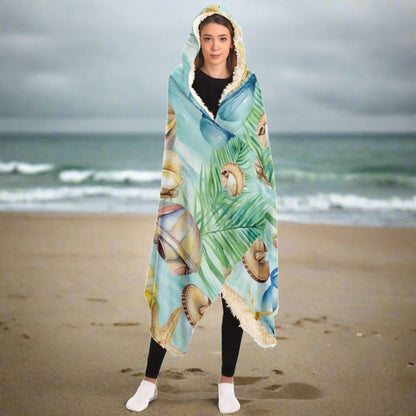 Person standing on a beach in a hooded blanket with a light aqua background with seashells and leaves with blue text that says Chill at the Beach.