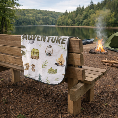 Camping scene with 'ADVENTURE' outdoor blanket on a wooden bench by a lake.