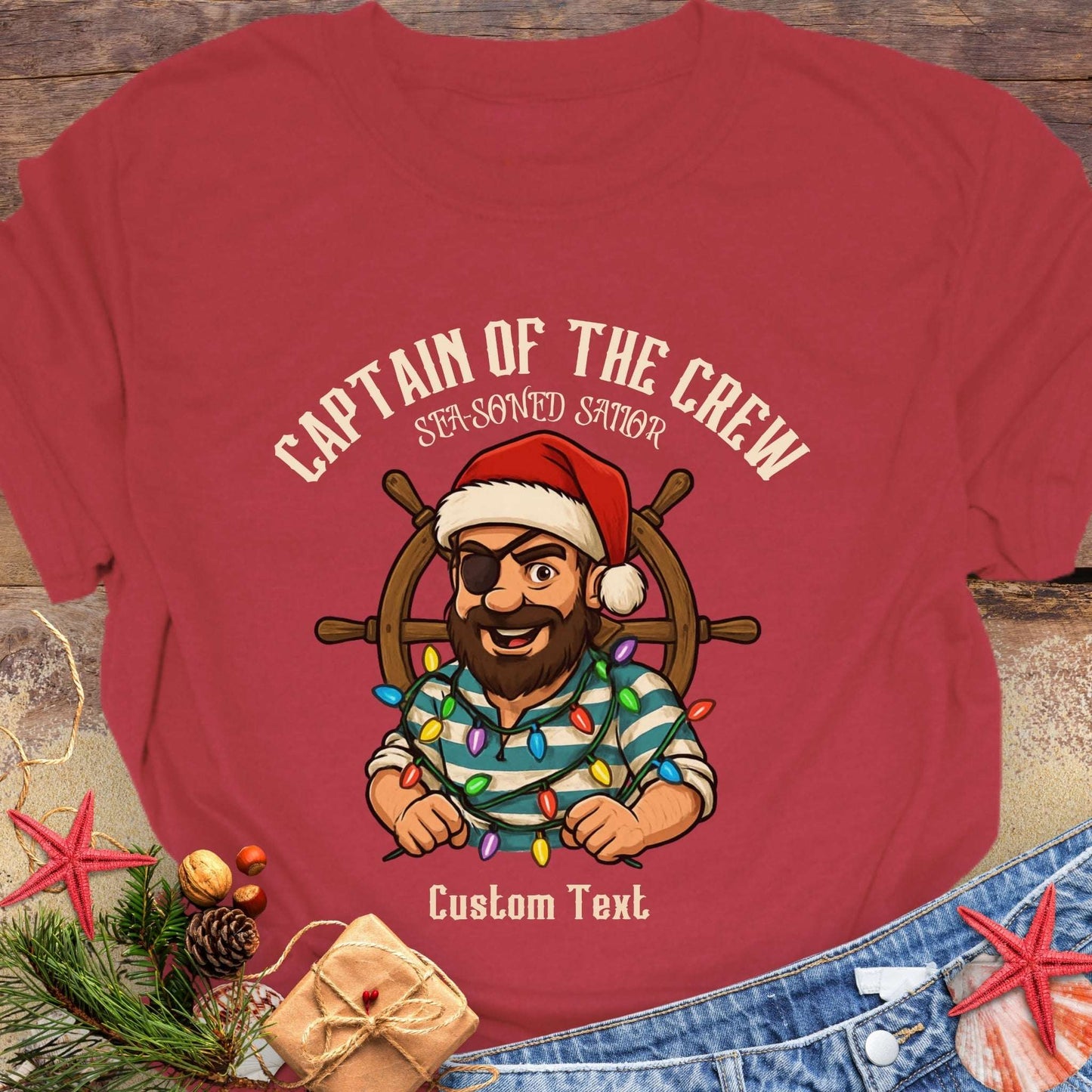 Red t-shirt with a cartoon character of a captain pirate wearing a Santa hat and holding Christmas lights, on a wooden surface with decorative items.