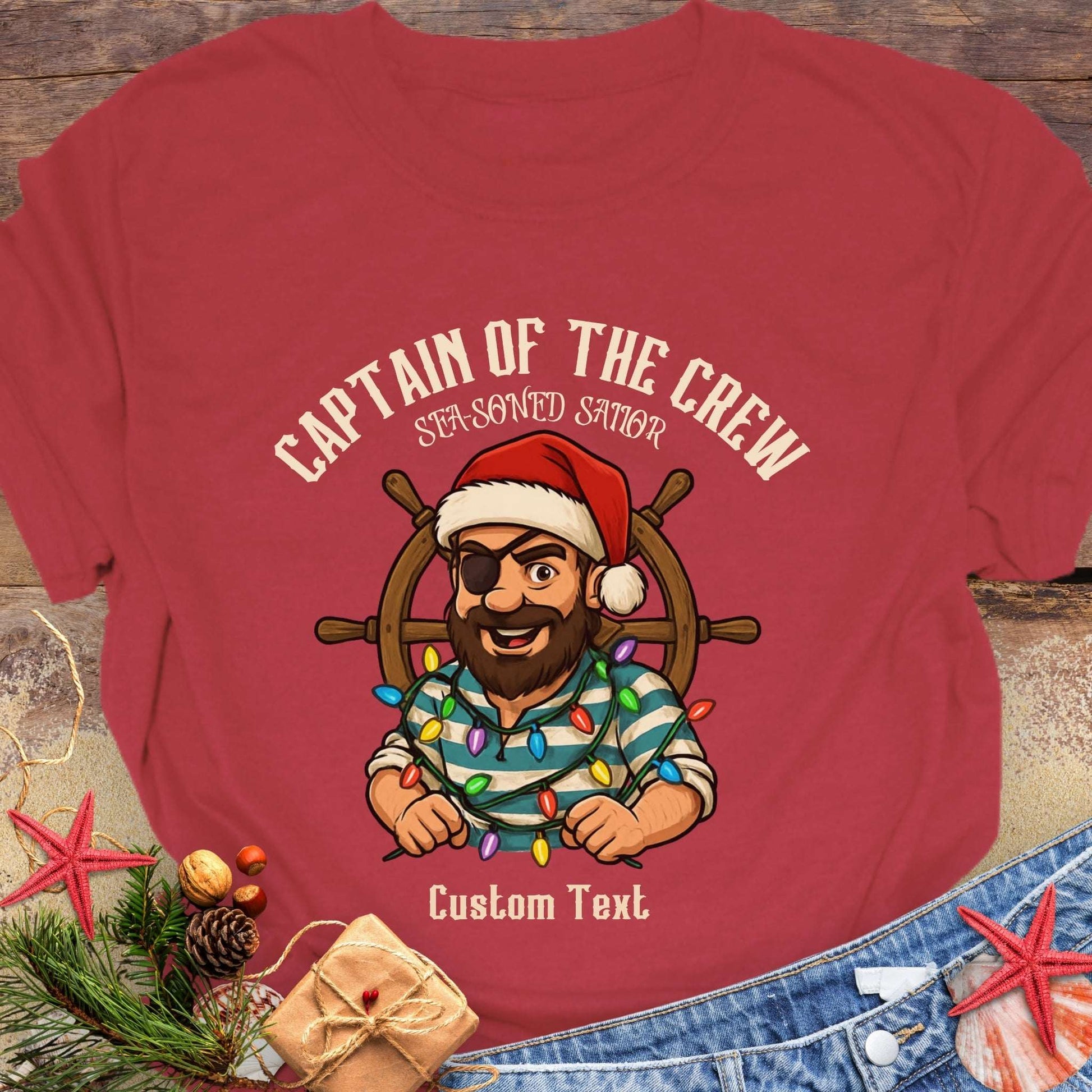 Red t-shirt with a cartoon character of a captain pirate wearing a Santa hat and holding Christmas lights, on a wooden surface with decorative items.