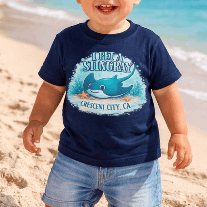 Child wearing a navy blue t-shirt with a graphic design of a stingray underwater and text I Pet a Stingray Crescent City, CA.