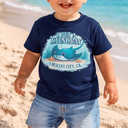 Child wearing a navy blue t-shirt with a graphic design of a stingray underwater and text I Pet a Stingray Crescent City, CA.