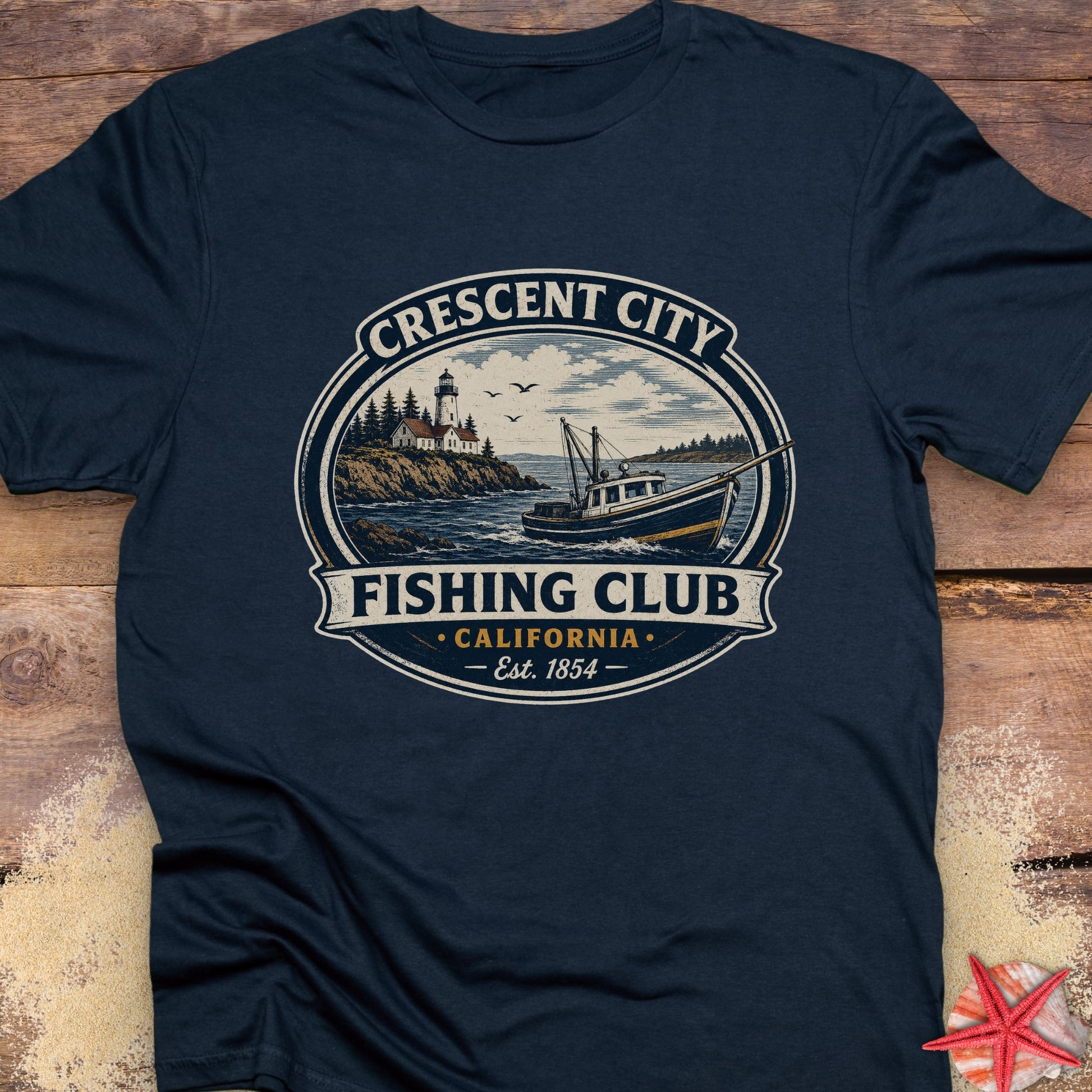 A navy blue short-sleeve t-shirt laid flat on a rustic wooden surface, featuring a large circular vintage-style graphic on the chest that reads 'Crescent City Fishing Club California Est. 1854' with an illustration of a lighthouse, boats, and water.