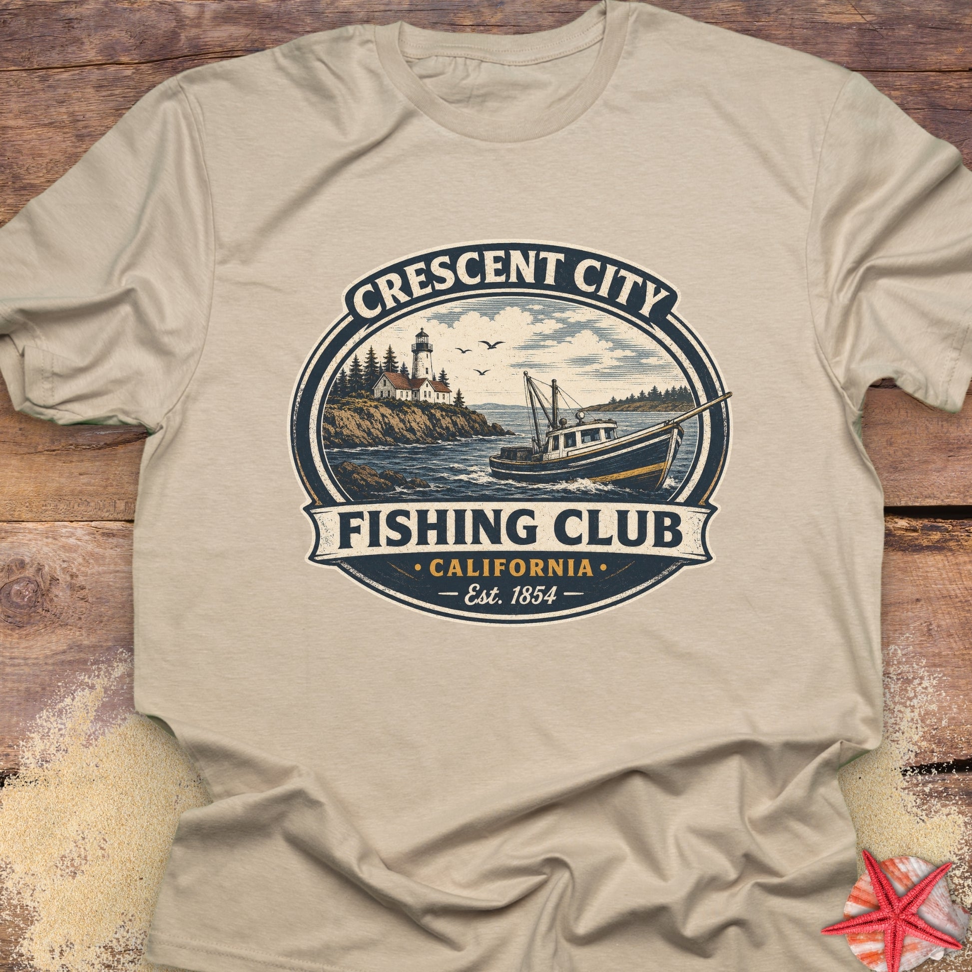 A beige crewneck t-shirt laid flat on a rustic wooden surface, featuring a vintage-style circular graphic for 'Crescent City Fishing Club, California' with an illustration of a fishing boat, lighthouse, and ocean.