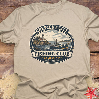 A beige crewneck t-shirt laid flat on a rustic wooden surface, featuring a vintage-style circular graphic for 'Crescent City Fishing Club, California' with an illustration of a fishing boat, lighthouse, and ocean.