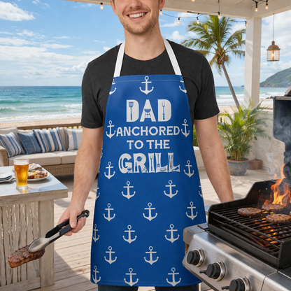 Man grilling outdoors wearing a blue apron with 'Dad Anchored to the Grill' text and anchor pattern.