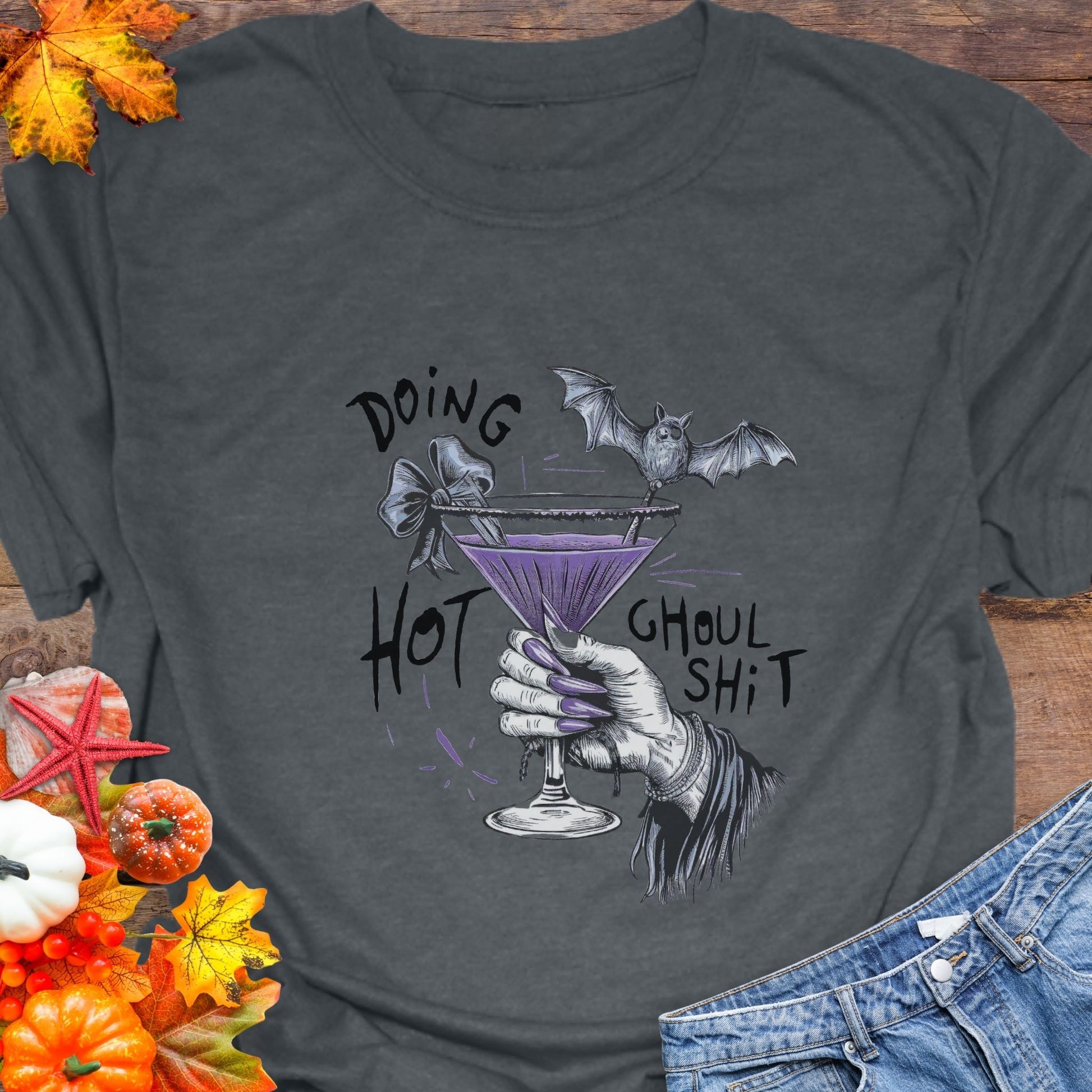 Gray t-shirt with a graphic of a hand holding a purple cocktail with a bat, text 'Doing Hot Ghoul Shit', on a wooden surface with Halloween decorations.