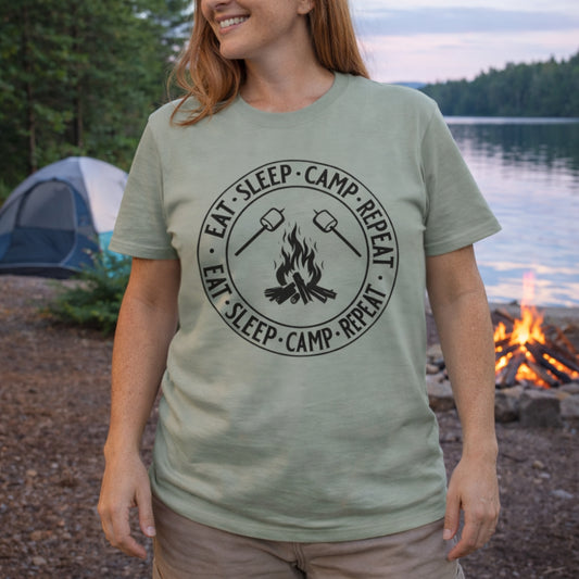 Person wearing a t-shirt with 'Eat Sleep Camp Repeat' design by a lake and campfire.