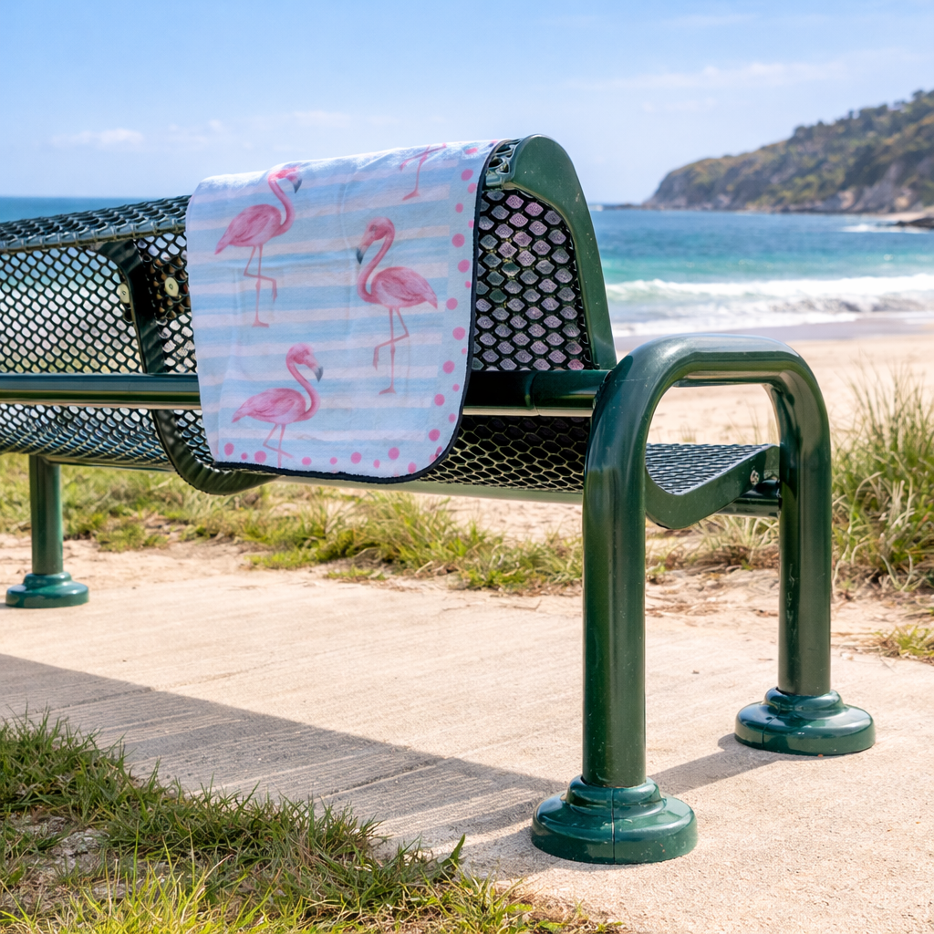 Green bench with a flamingo-patterned beach blanket draped over it by the beach.