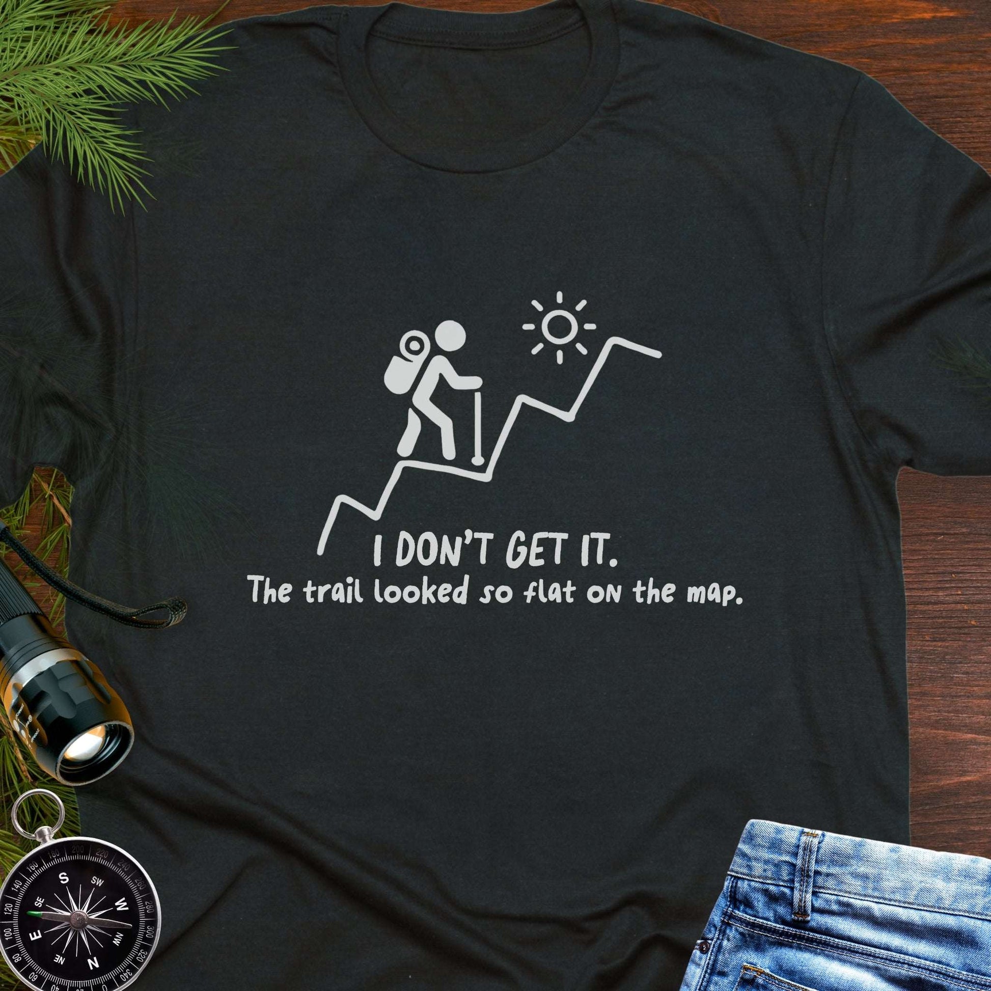 Black hiking t-shirt flat lay with white graphic of a backpacker climbing a steep trail and text: “I DON’T GET IT. The trail looked so flat on the map.”