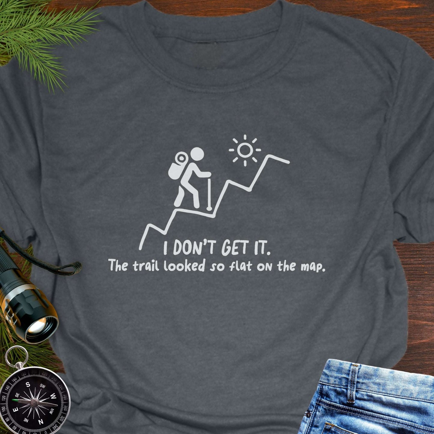 Dark Gray hiking t-shirt flat lay with white graphic of a backpacker climbing a steep trail and text: “I DON’T GET IT. The trail looked so flat on the map.”