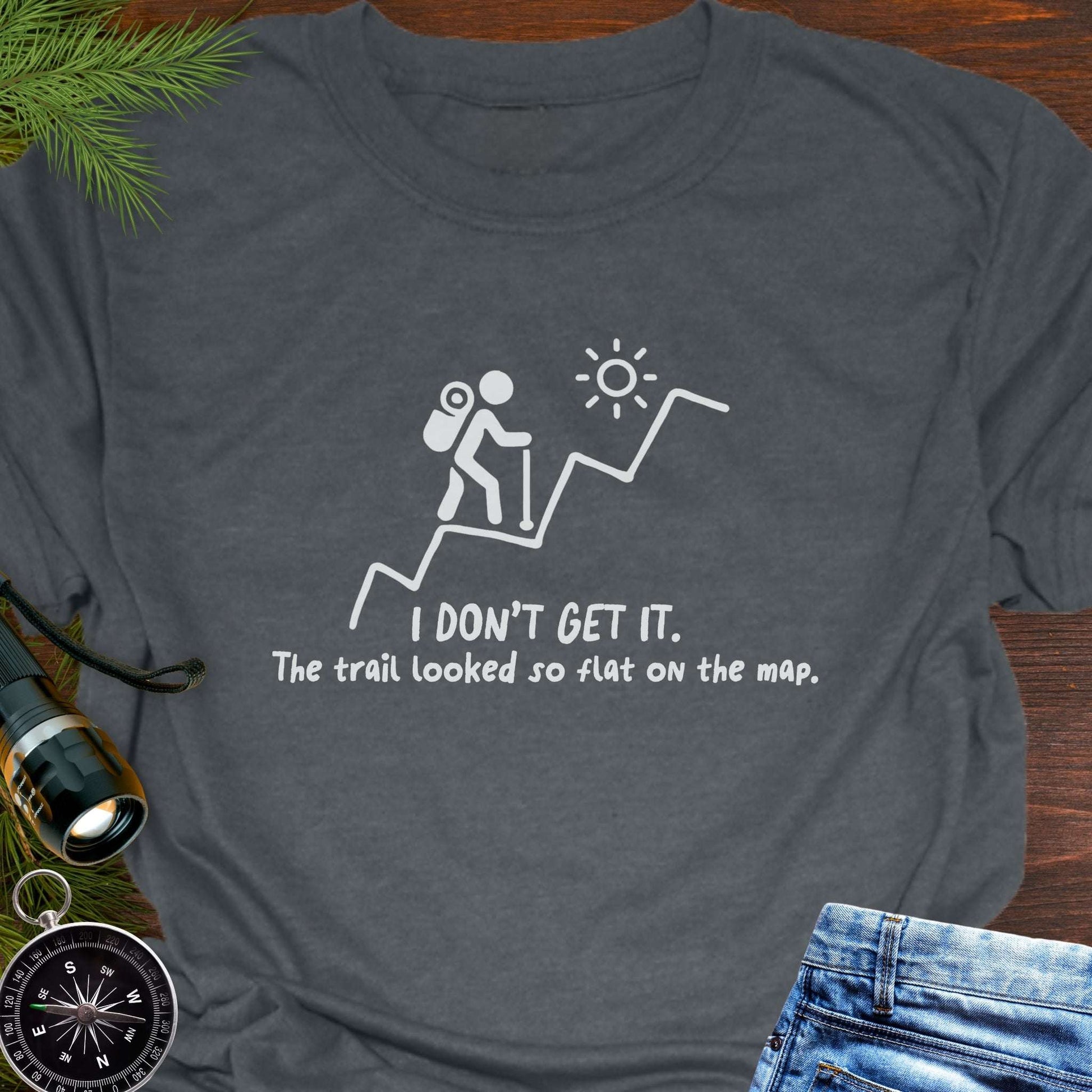 Dark Gray hiking t-shirt flat lay with white graphic of a backpacker climbing a steep trail and text: “I DON’T GET IT. The trail looked so flat on the map.”