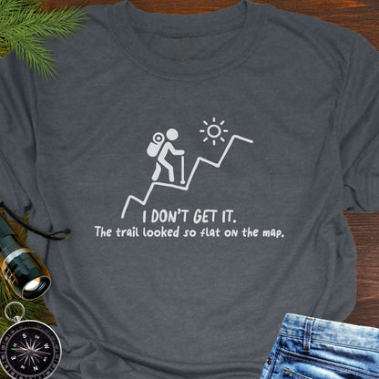 Dark Gray hiking t-shirt flat lay with white graphic of a backpacker climbing a steep trail and text: “I DON’T GET IT. The trail looked so flat on the map.”