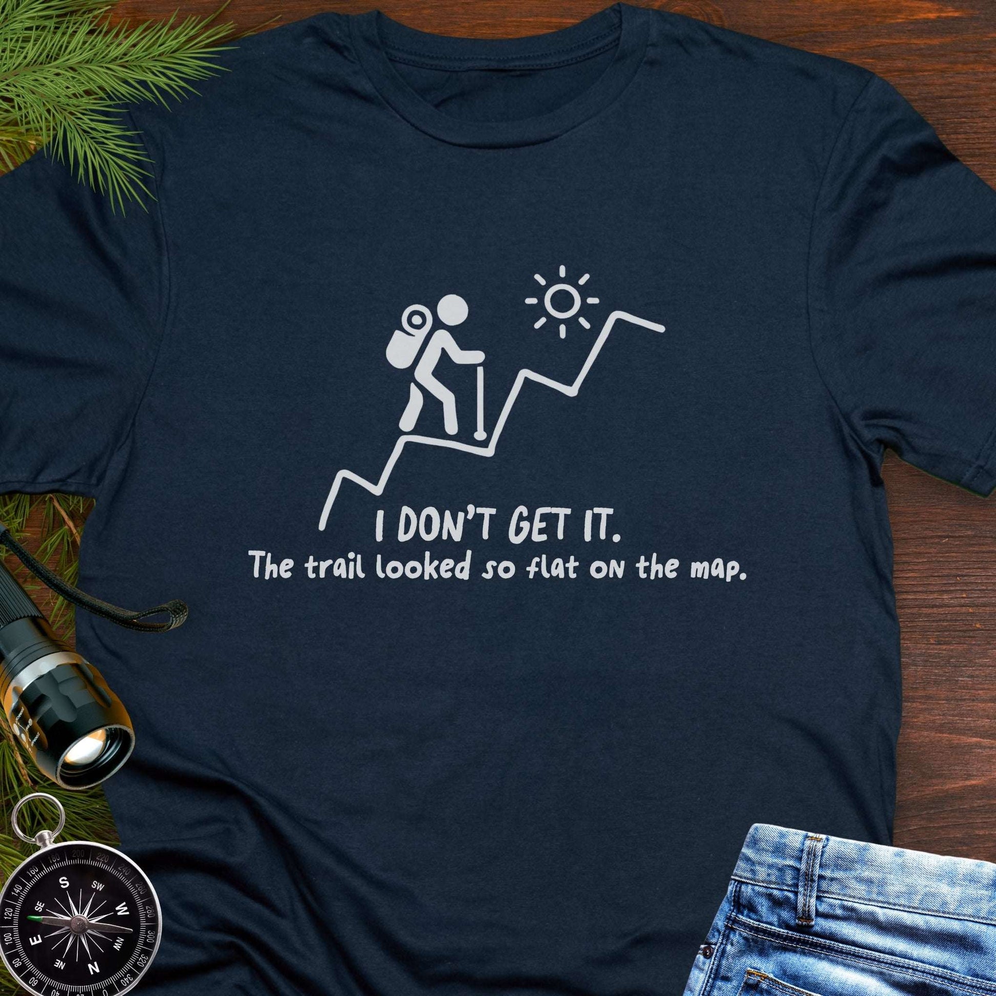 Navy blue hiking t-shirt flat lay with white graphic of a backpacker climbing a steep trail and text: “I DON’T GET IT. The trail looked so flat on the map.”