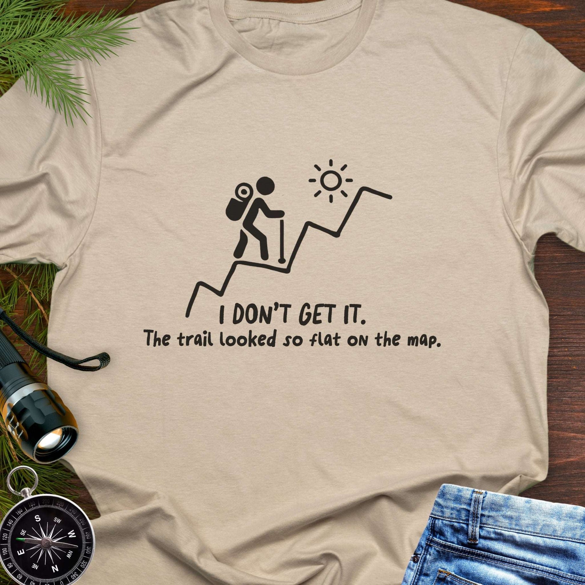 Beige hiking t-shirt flat lay with black graphic of a backpacker climbing a steep trail and text: “I DON’T GET IT. The trail looked so flat on the map.”