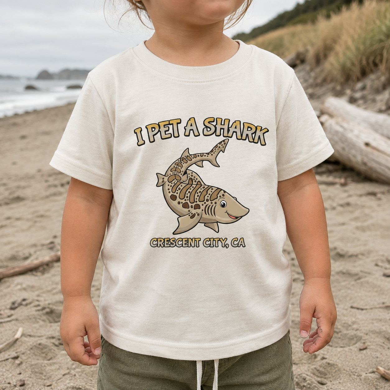 A toddler wearing a natural beige graphic t-shirt with a cartoon shark and the text 'I PET A SHARK' and 'CRESCENT CITY, CA' on a sandy beach.