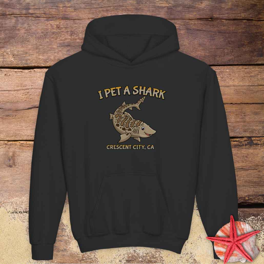 A black pullover hoodie with a graphic print on the chest reading 'I PET A SHARK' and 'CRESCENT CITY, CA' with a shark illustration.