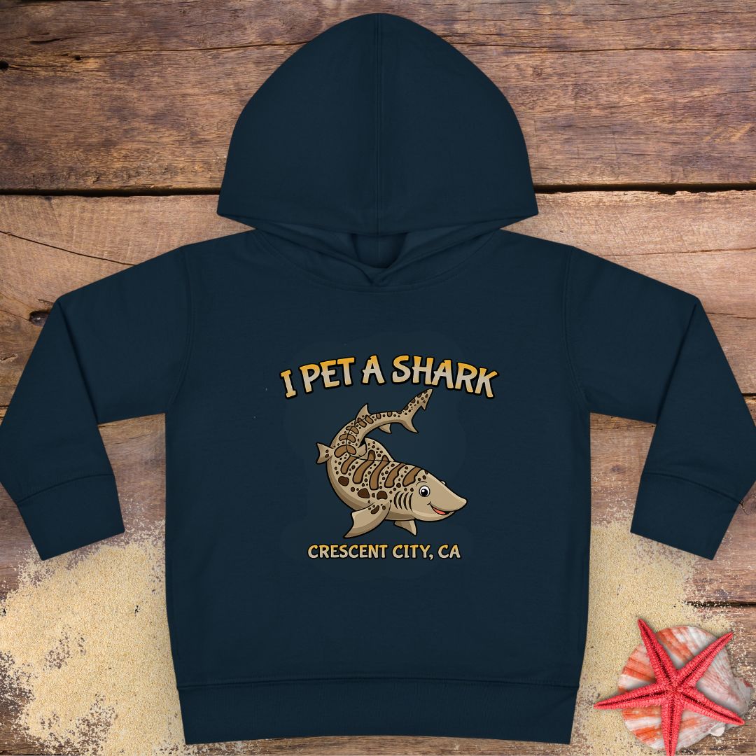 A navy blue Toddler hoodie featuring a smiling cartoon shark with the phrase "I Pet a Shark" and "Crescent City, CA," perfect for little shark lovers and ocean adventures.