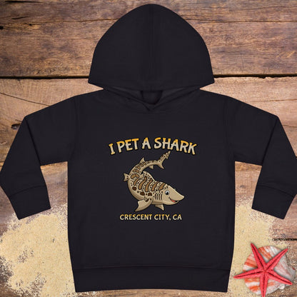 A black Toddler hoodie featuring a smiling cartoon shark with the phrase "I Pet a Shark" and "Crescent City, CA," perfect for little shark lovers and ocean adventures.