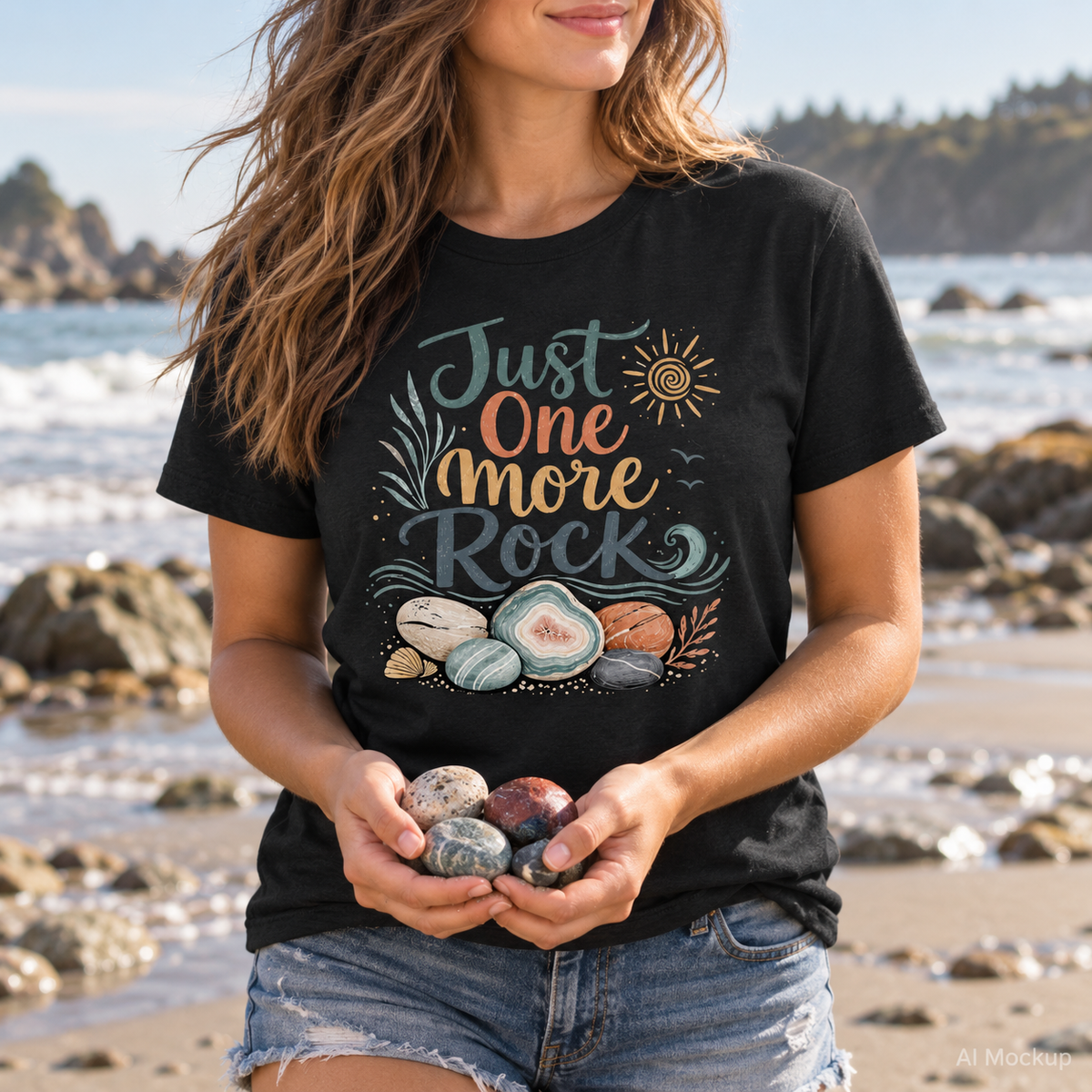 Woman wearing a black "Just One More Rock" graphic t-shirt featuring colorful beach stones and agates while holding smooth rocks on a sandy beach.