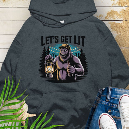 Dark Heather funny Sasquatch camping unisex hoodie with relaxed outdoor party vibe that says Let's Get Lit featuring a sasquatch wearing a ball cap holding a camping lantern and a bottle of beer with the forest in a forest camp scene.