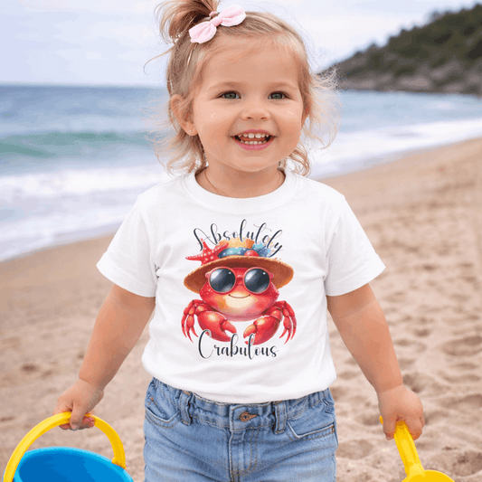 Child on a beach wearing a white t-shirt with a colorful crab wearing a fancy hat design and text 'Absolutely Crabulous'.