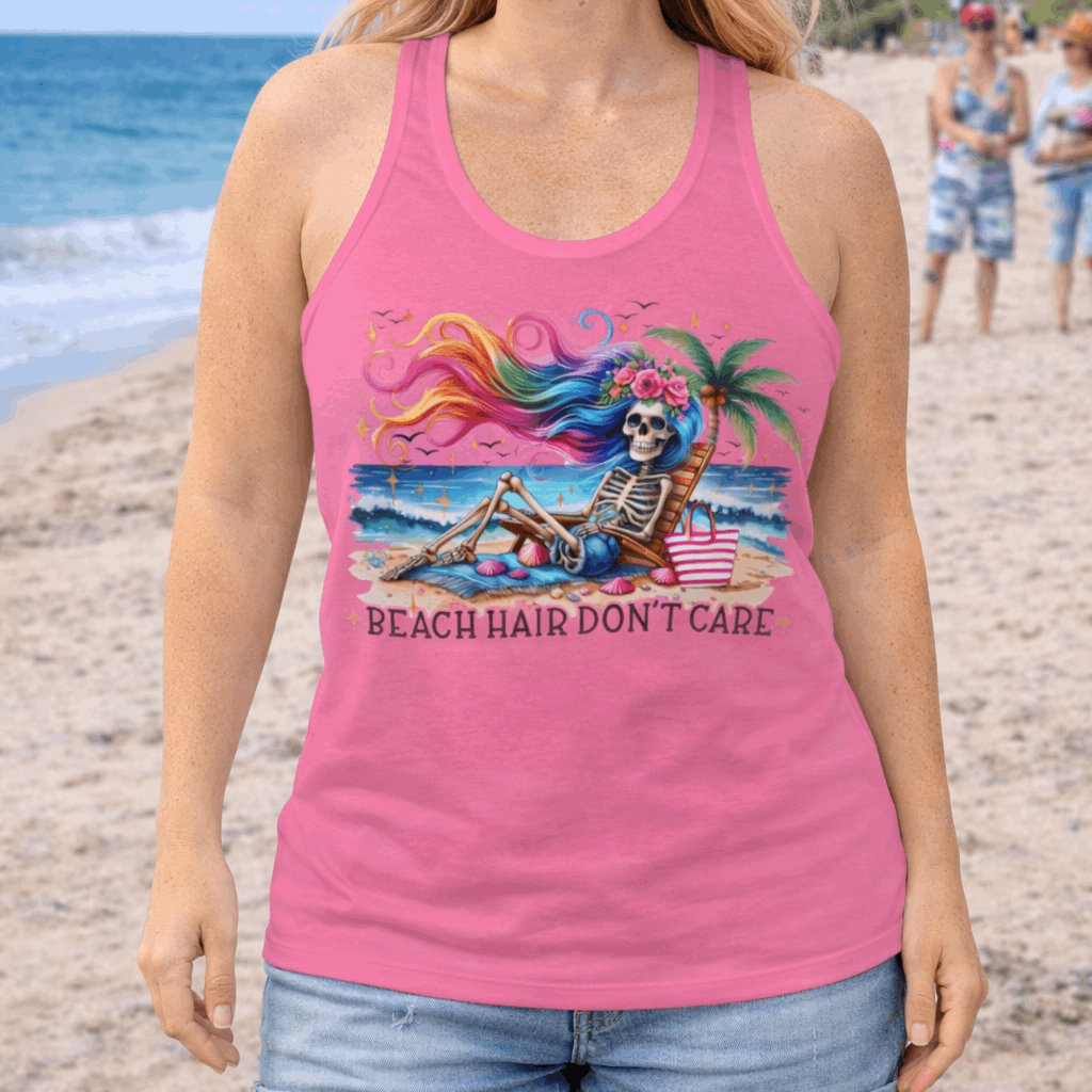 Woman wearing a pink Racerback Tank Top with a skeleton with rainbow hair flowing in the wind laying on a beach chair at the ocean that says Beach Hair Don't Care.