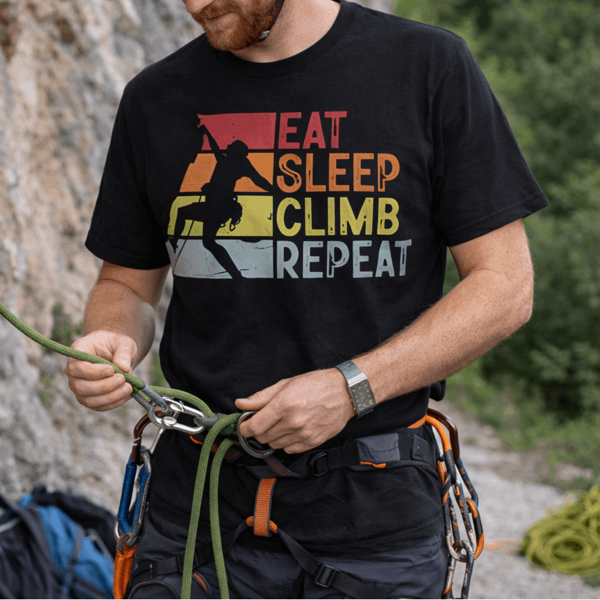 Man preparing for climbing with 'Eat Sleep Climb Repeat' t-shirt against a rock wall.