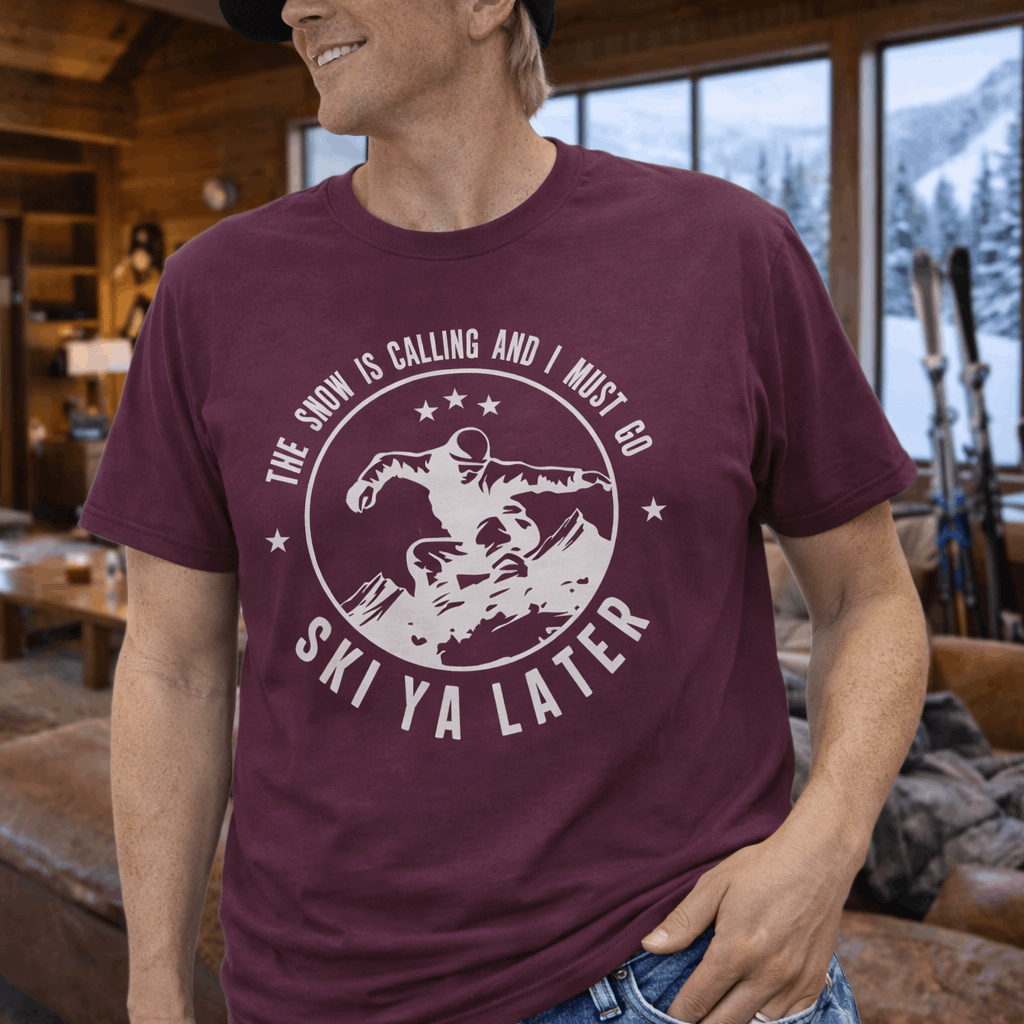 Man wearing a maroon t-shirt with a skiing design and text the snow is calling and I must go ski ya later.
