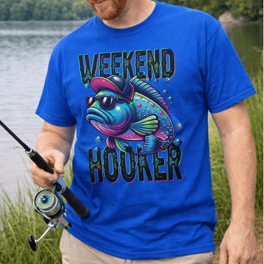 Man fishing by a lake wearing a blue t-shirt with a colorful fish graphic and text Weekend Hooker.