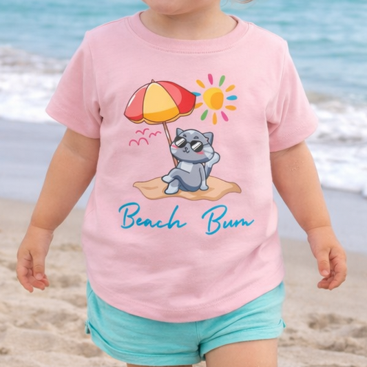 Child wearing a pink toddler t-shirt with vibrant playful summer design of a cute cat wearing sunglasses laying on the beach under an umbrella with text Beach Bum. 