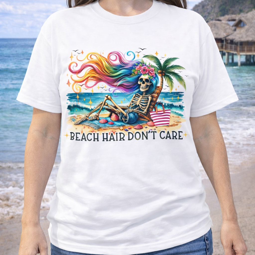 A woman wearing a white t-shirt with a skeleton with rainbow hair flowing in the wind laying on a beach chair at the ocean that says Beach Hair Don't Care.