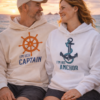 Couple wearing matching hoodies with 'I'm her Captain' and 'I'm his Anchor' designs, sitting on a beach at sunset.
