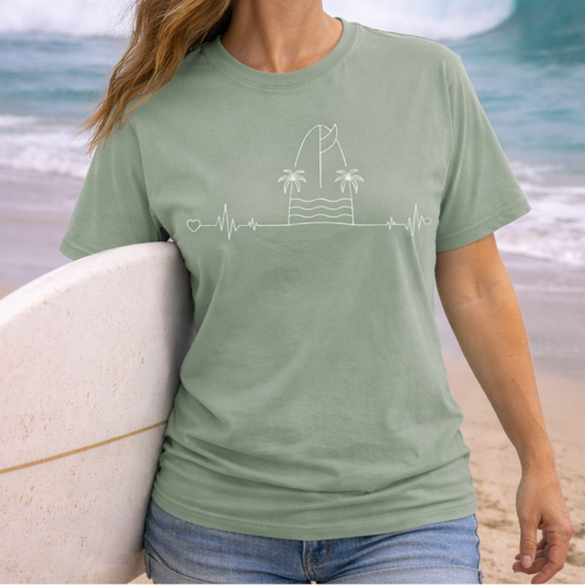Woman wearing a green t-shirt with a beach heartline design with a surfboard and palm trees. 