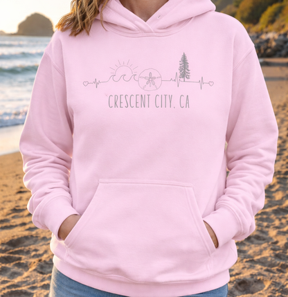 Person wearing a pink hoodie with a heartline with waves, sand dollar, redwood tree and sun with text 'Crescent City, CA'.