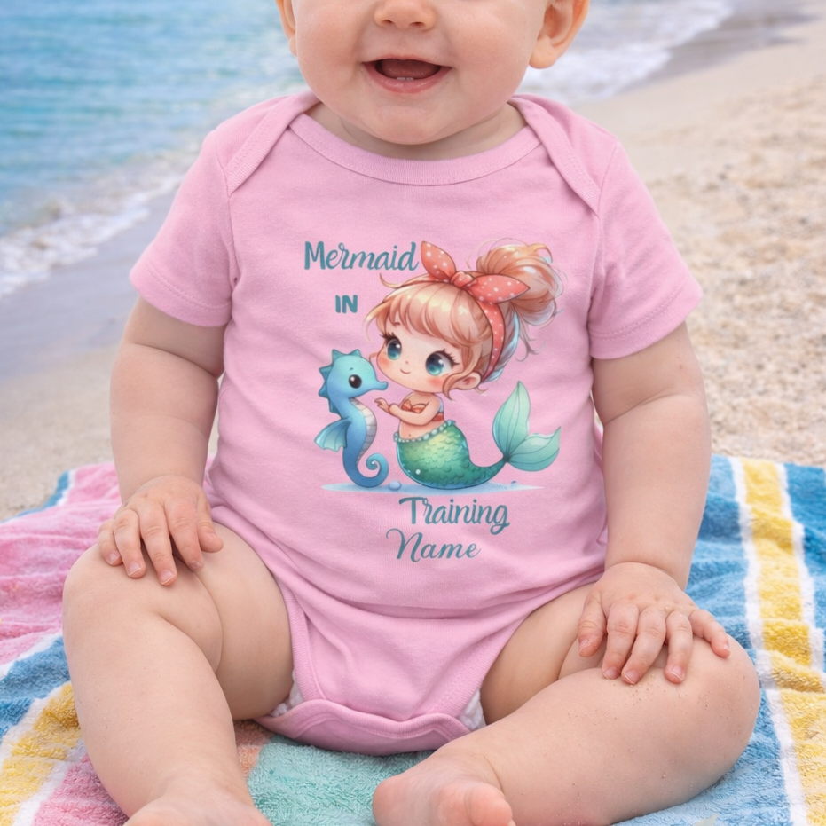 Baby in pink 'Mermaid in Training' with personalization onesie on a beach.