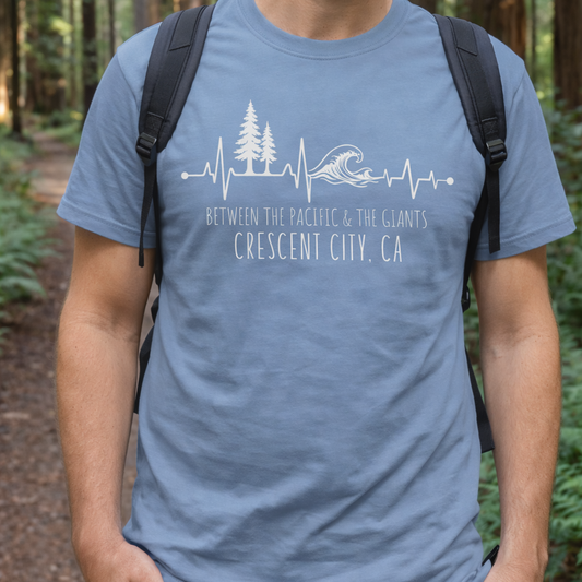 Man wearing a blue t-shirt with a nature-themed heartline design of redwoods and ocean waves with text Between the Pacific & The Giants Crescent City, CA.