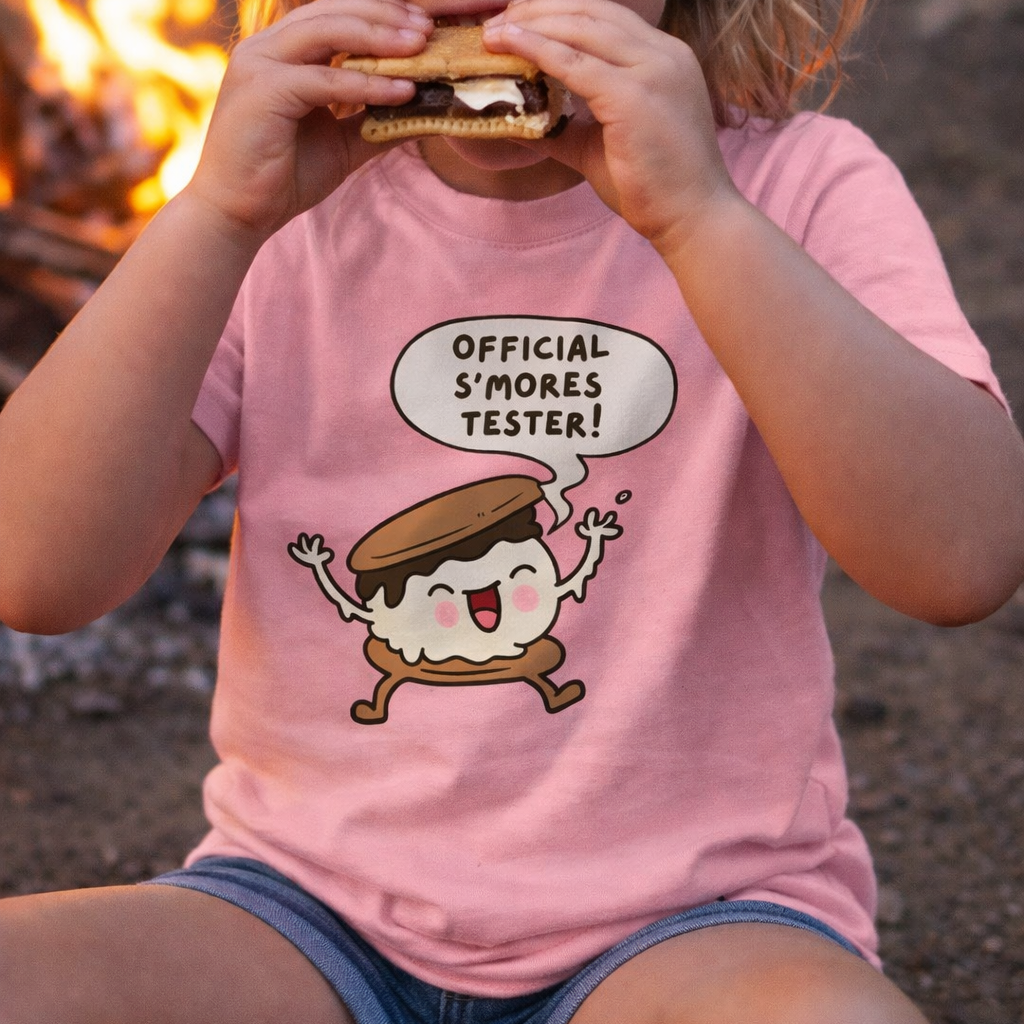 Child eating s'mores with a pink toddler t-shirt featuring a cartoon s'more character and text Official S'mores Tester.