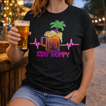 Person wearing a black unisex t-shirt with a neon-style graphic of a beer mug, palm tree, and heartbeat line, featuring the phrase “Stay Hoppy” in bold pink and purple.