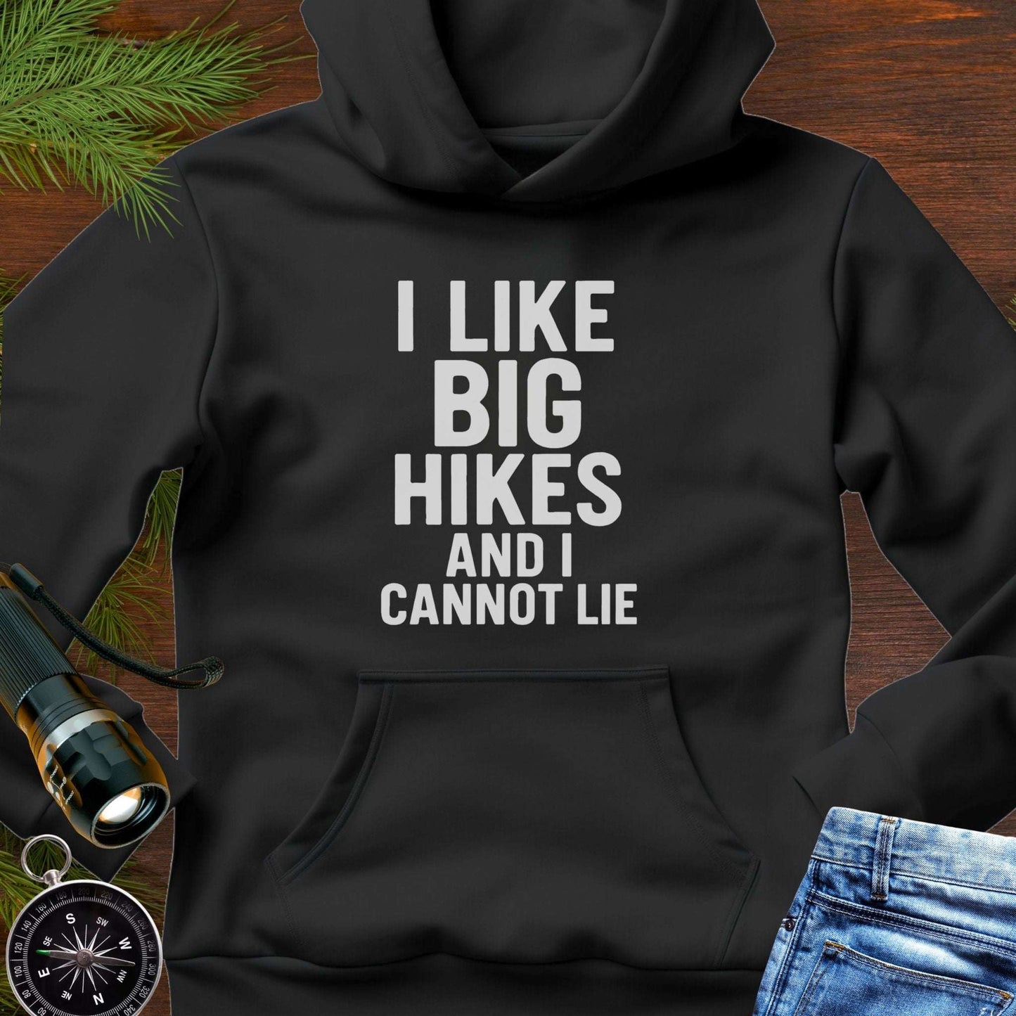 Black hoodie with 'I LIKE BIG HIKES AND I CANNOT LIE' text on a wooden surface with hiking accessories.