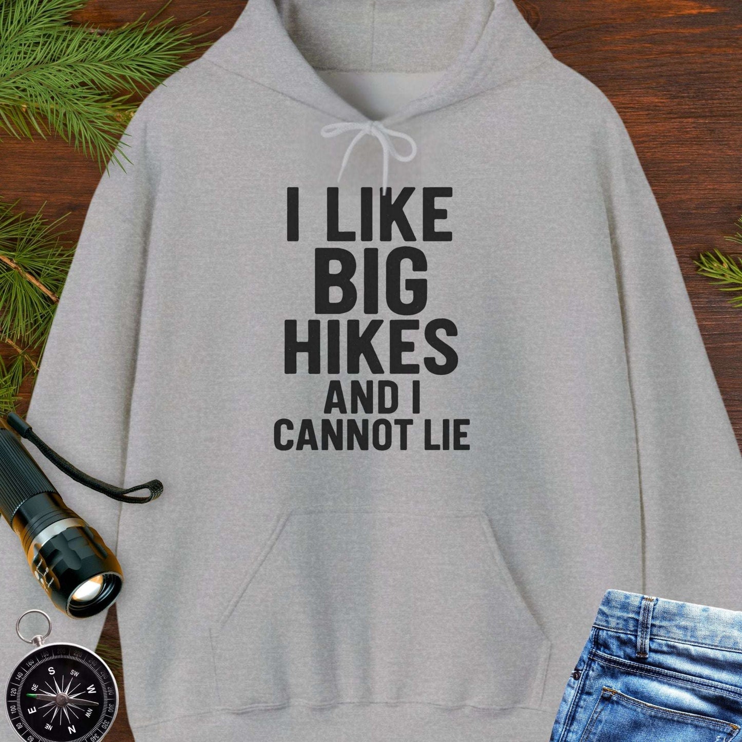 Gray hoodie with 'I LIKE BIG HIKES AND I CANNOT LIE' text, surrounded by hiking gear on a wooden surface.