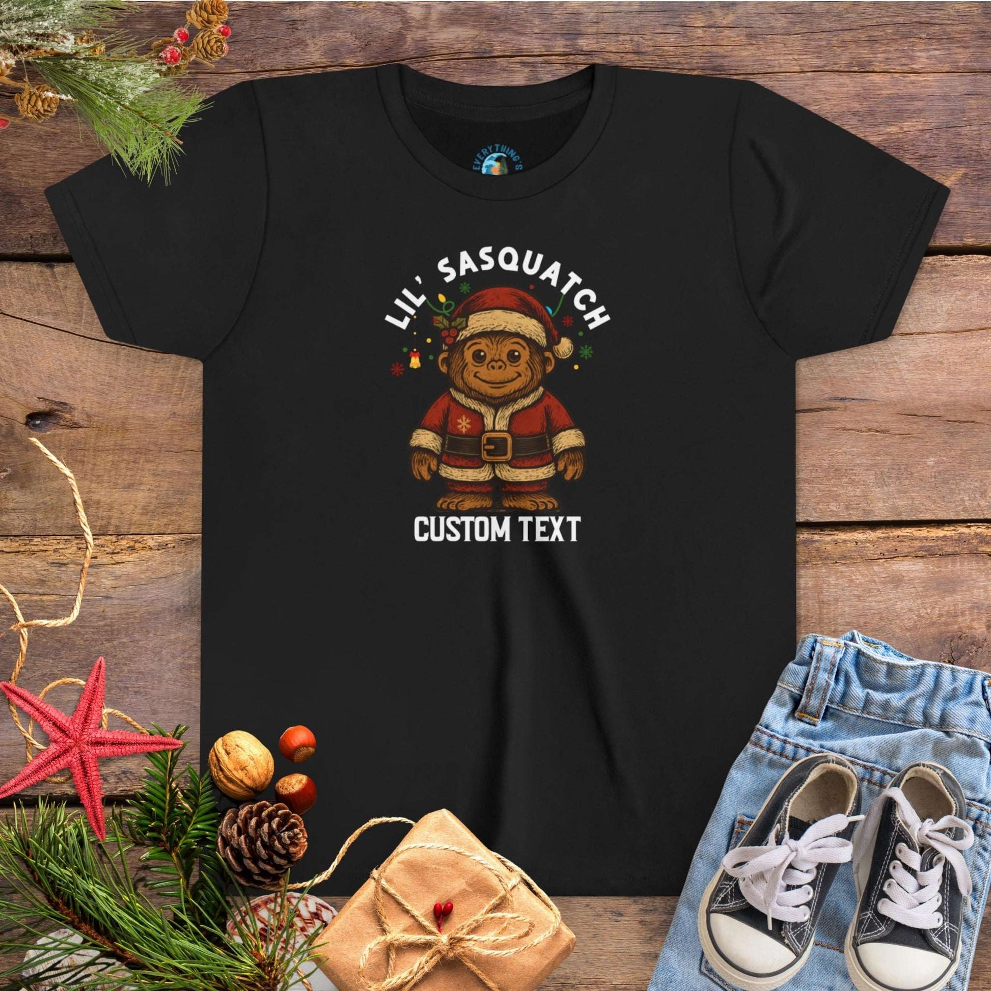 Black t-shirt with Christmas-themed graphic and 'Lil Sasquatch' text on a wooden surface with festive decorations.