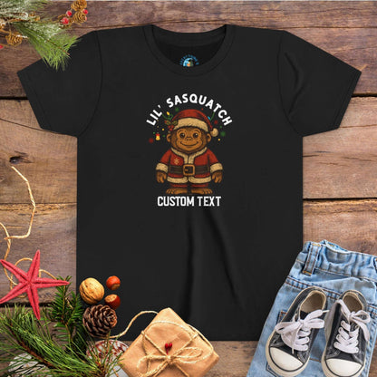 Black t-shirt with Christmas-themed graphic and 'Lil Sasquatch' text on a wooden surface with festive decorations.